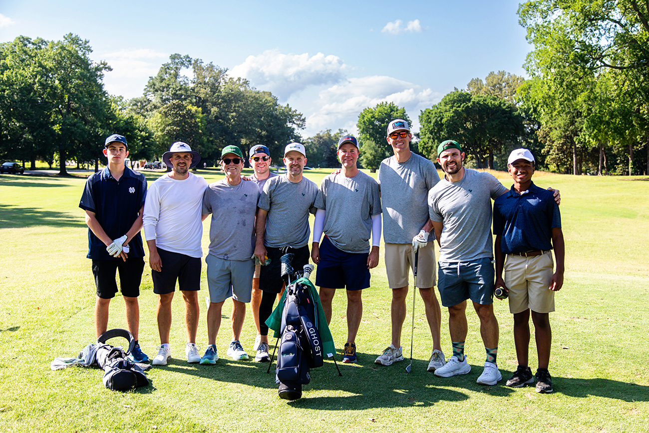 A group of golfers in gray t-shirts stand on a golf course in the afternoon