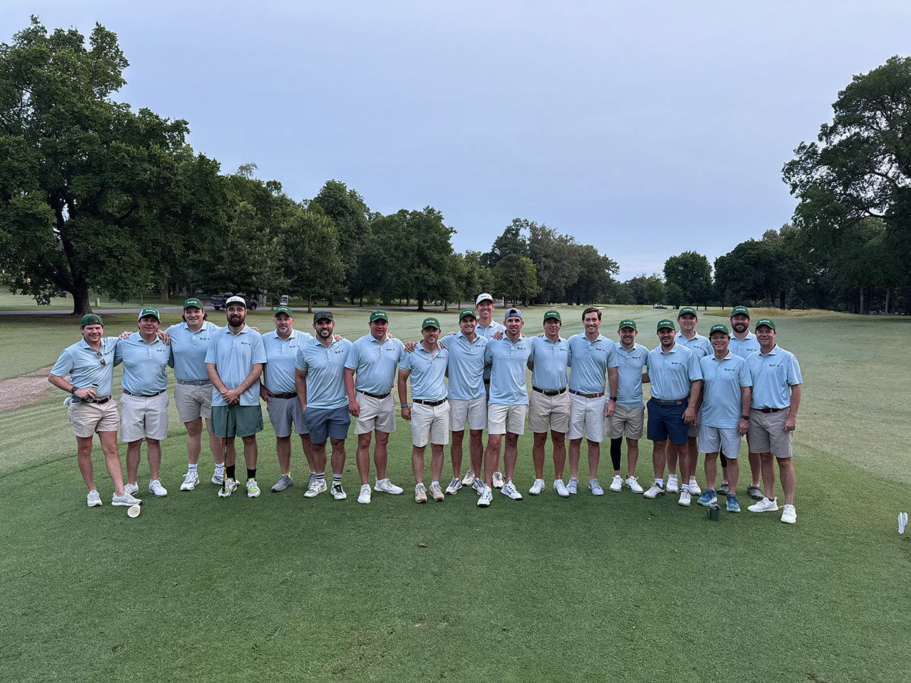 A group of golfers wearing light blue shirts stand on a golf course